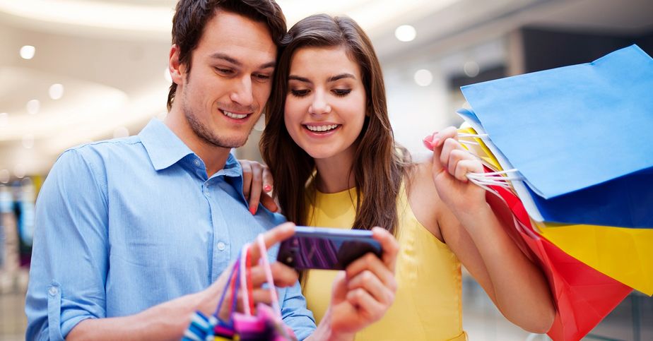 Dónde comprar iPhone y celulares en Quito | Centro Comercial Caracol Pareja joven revisando un celular mientras sostiene bolsas de compras dentro de un centro comercial.